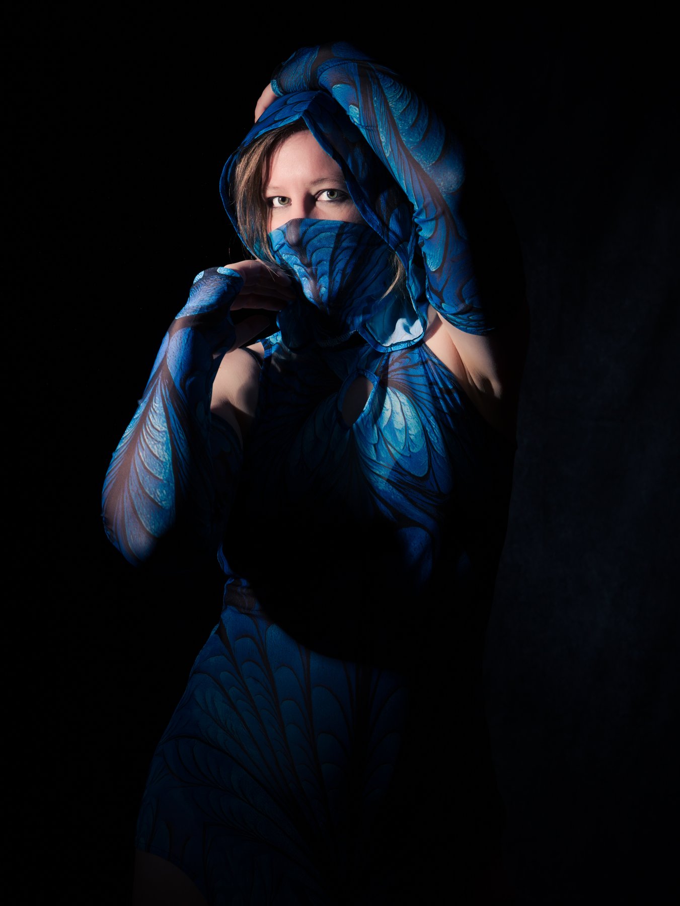 a woman wrapped in blue butterfly-patterned fabric veiling her lower face against a black background
