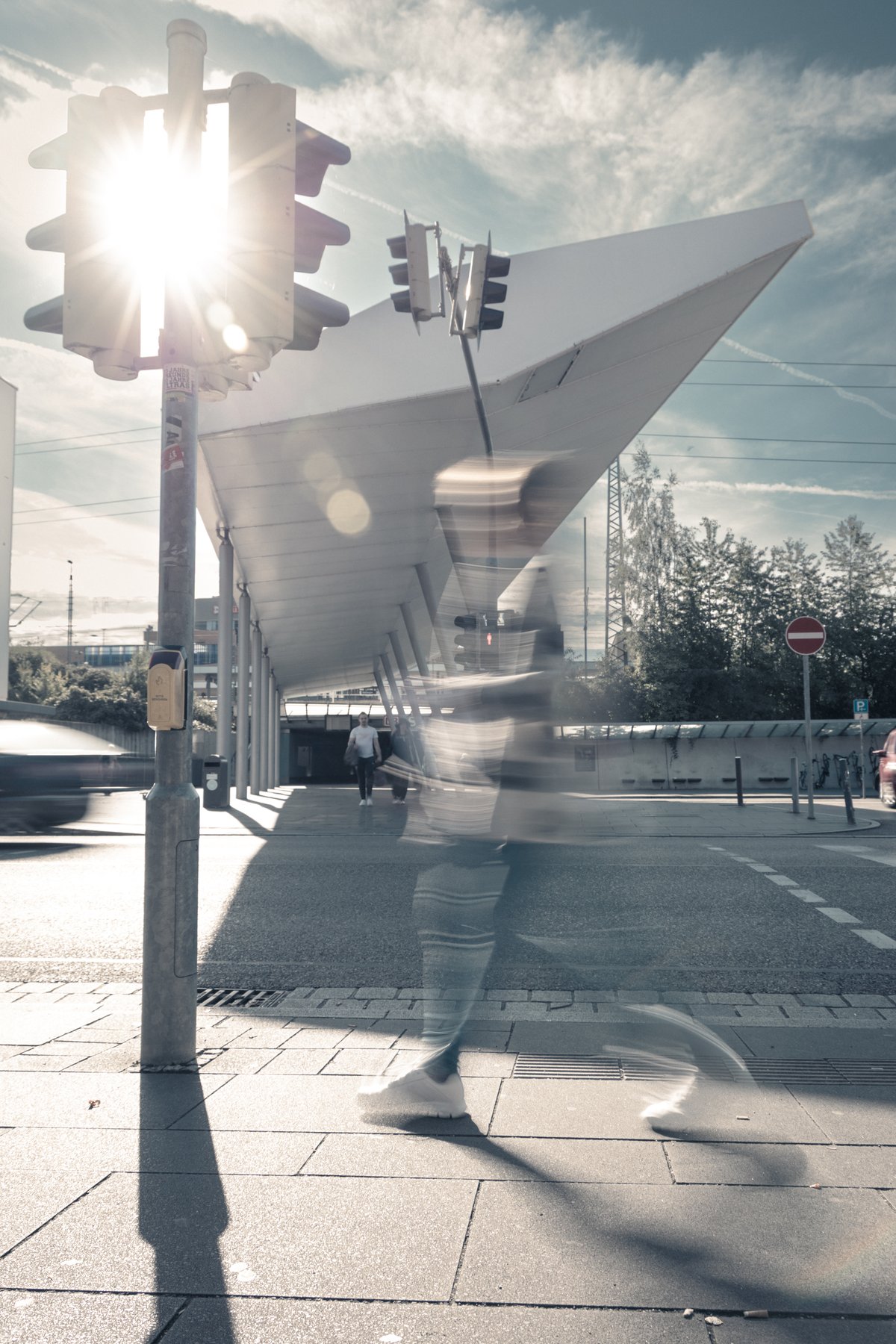 a motion-blurred pedestrian crossing beneath an angular canopy, sun flaring behind a traffic light pole