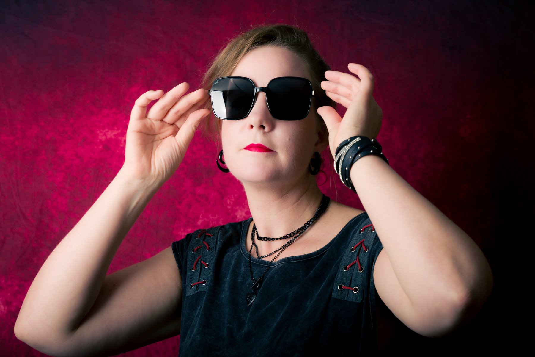 a woman with red lipstick adjusting large square sunglasses against a mottled red studio backdrop