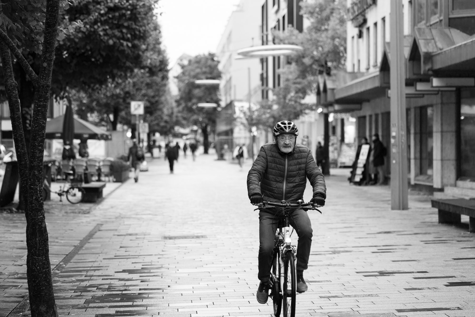 an older man in a helmet cycling down a wet pedestrian shopping street, black and white