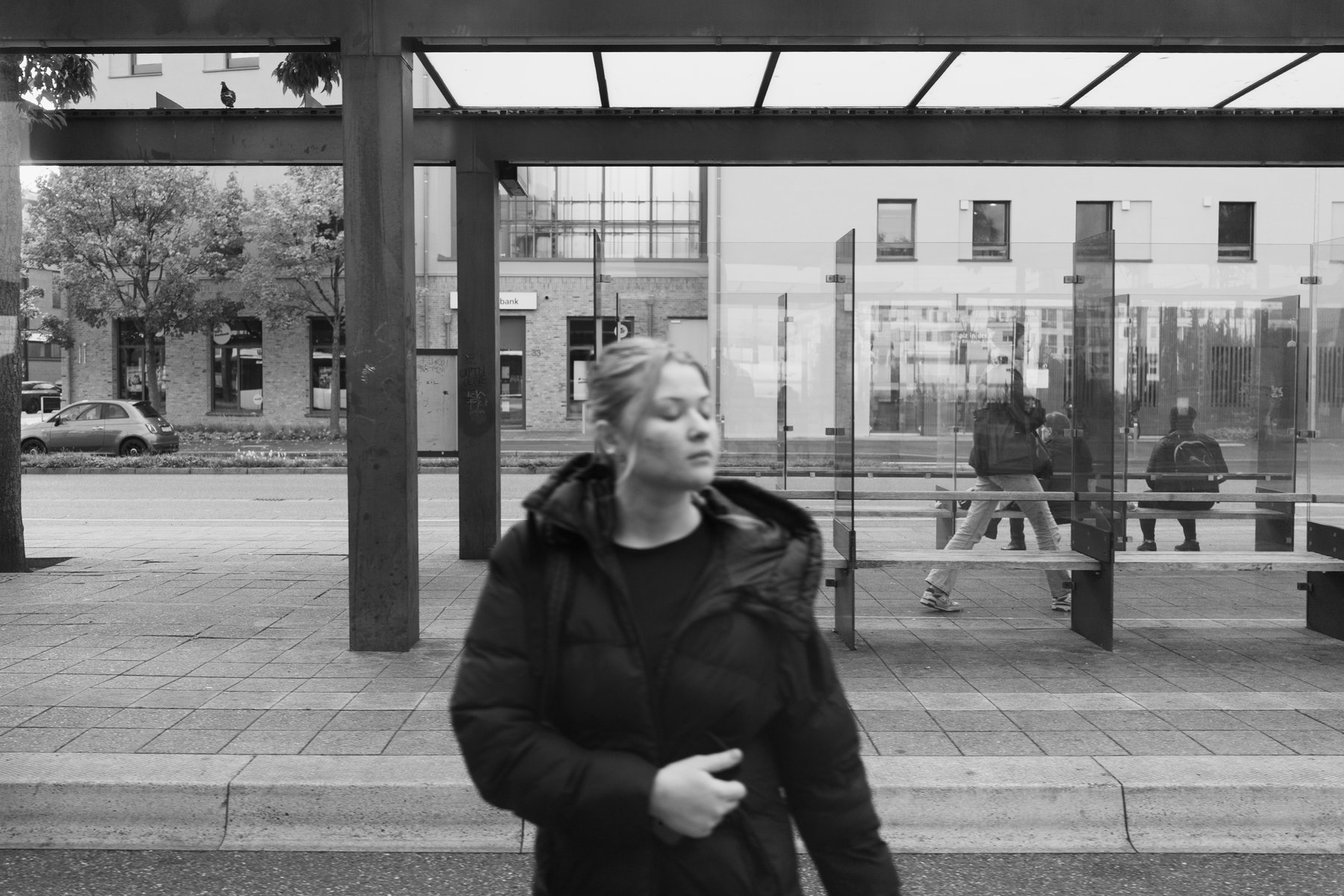 a woman in a dark coat walking past a glass tram shelter with seated commuters behind