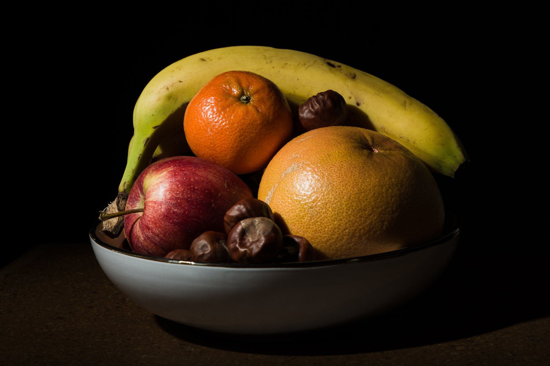 fruit bowl with banana, orange, apple, grapefruit, and chestnuts against dark background