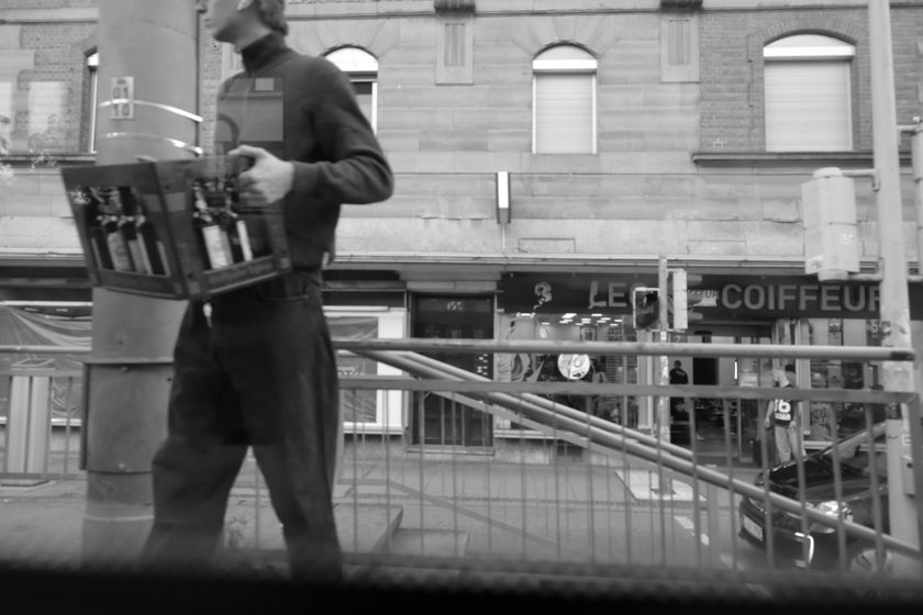 a man carrying a crate of bottles past street railings and a hairdresser storefront, black and white