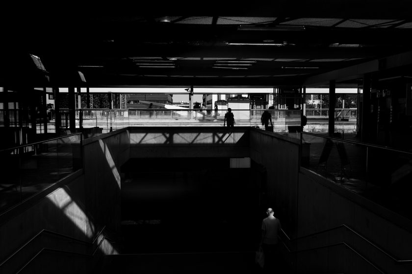silhouetted figures in a dark station concourse, sunlight cutting across the walkway above a stairwell
