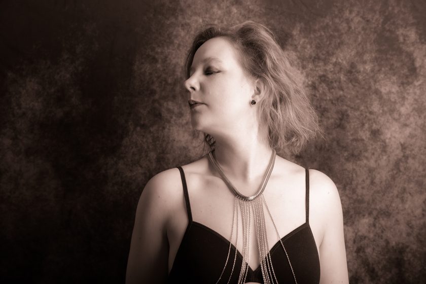 a woman in a black top and chain necklace, eyes closed, against a mottled sepia backdrop